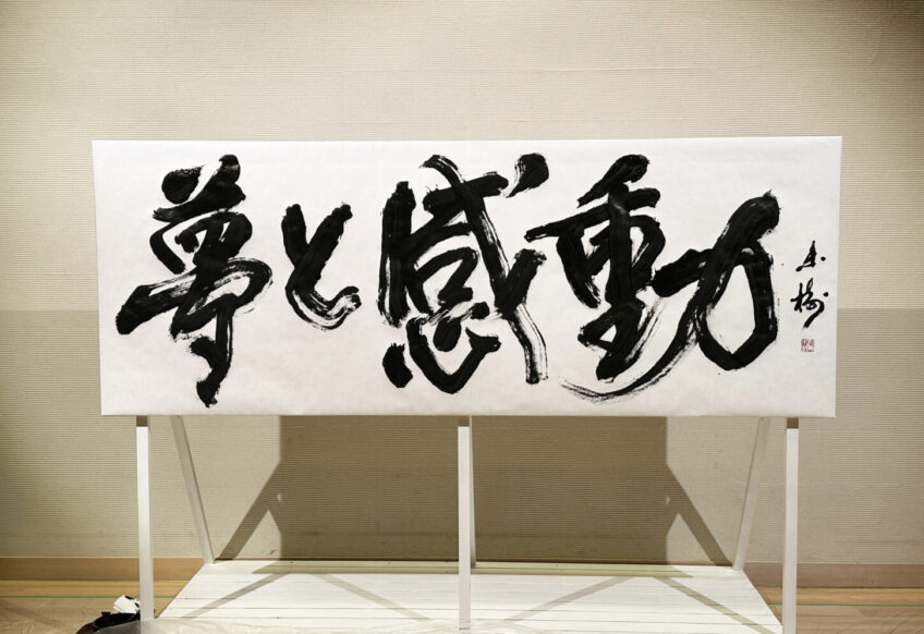 書家・根本みきが「DIグループ安全衛生協議会 創立20周年記念祝賀会」で、DIグループ代表・小板橋社長のモットー「夢と感動」を力強く揮毫する書道パフォーマンスを披露。参加者の夢が実り、感動が広がるよう願いを込めた作品。